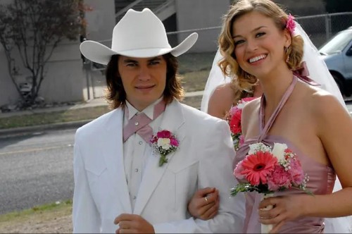 Tim Riggins wearing a white suit and white cowboy hat arm in arm with Tyra, wearing a pink bridesmaid dress and holding a bouquet