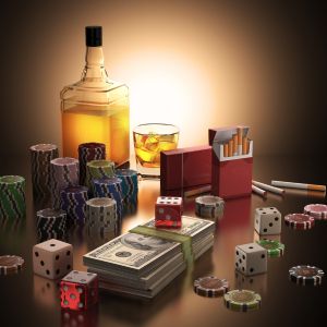 A low-lit photo of a collection of alcohol, poker chips, dice, a stack of money, and a pack of cigarettes.